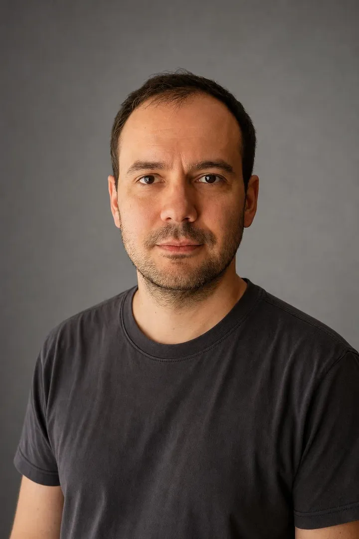 The same man in a studio headshot in his own grey t shirt, grey backdrop.
